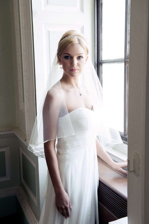 Bride at window wearing two layer elbow length single layer veil with ribbon edging